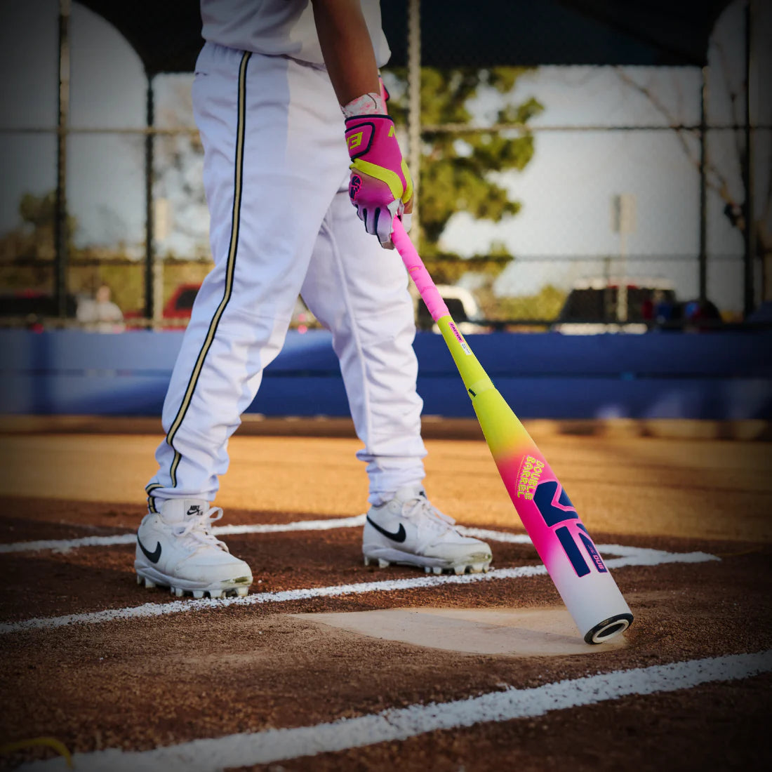 A youth baseball player in white pants and a white and pink baseball bat with the brand 'Easton' visible, standing in a batters box with pink gloves.